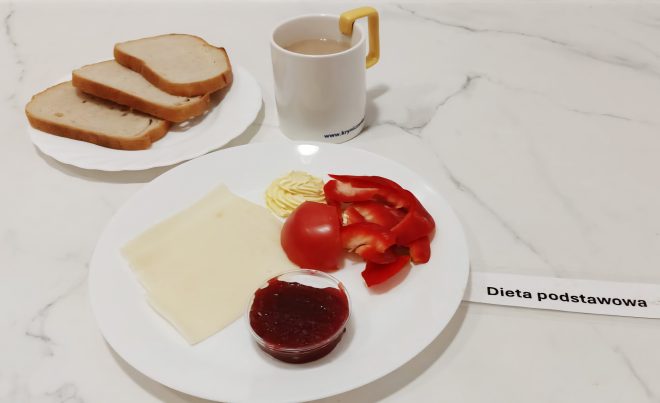 Śniadanie w diecie podstawowej. Na talerzu pieczywo pszenne, plasterki sera, masło, papryka i pomidor oraz porcja dżemu. Obok kubek z kawą z mlekiem