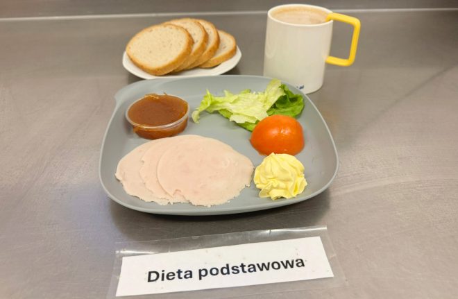 Na stalowym blacie znajduje się śniadanie oznaczone etykietą „Dieta podstawowa”. Na szarym talerzu ułożono kilka plasterków chudej wędliny drobiowej, porcję sałaty, połówkę pomidora, odrobinę masła oraz mały kubeczek z dżemem. Z tyłu talerza leży kilka kromek jasnego pieczywa. Obok stoi biały kubek z żółtym uchem, wypełniony napojem – prawdopodobnie kawą z mlekiem lub kakao