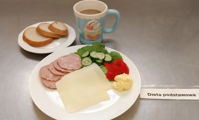 Śniadanie w diecie podstawowej. Na talerzu plasterki wędliny, ser żółty, masło, sałata, ogórek i papryka. Obok filiżanka z kawą z mlekiem i talerzyk z trzema kromkami białego pieczywa