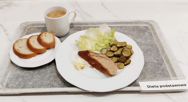 Śniadanie w diecie podstawowej: kromki chleba na małym talerzyku, kawa z mlekiem w białym kubku, a na dużym talerzu porcja pieczeni z masłem, sałatą i plasterkami ogórków kiszonych. Całość podana na tacy z kartką z napisem „Dieta podstawowa”