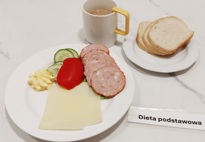 Na tacy podano śniadanie w diecie podstawowej. Na talerzu znajdują się plasterki kiełbasy, żółty ser, kawałek pomidora, plasterki świeżego ogórka oraz rozetka masła. Obok stoi kubek kawy z mlekiem i talerzyk z kilkoma kromkami jasnego pieczywa
