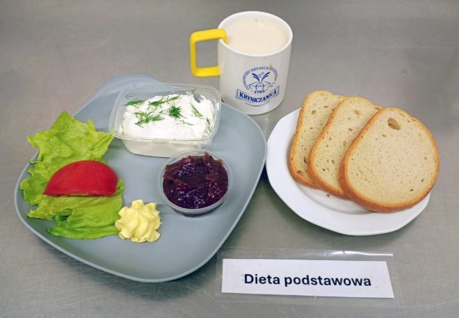 Śniadanie w diecie podstawowej. Na tacy podano trzy kromki chleba pszennego, porcję twarożku z koperkiem, dżem, liście sałaty z plastrem pomidora, porcję asła oraz kubek z napojem mlecznym. Z przodu widoczna kartka z napisem „Dieta podstawowa”.