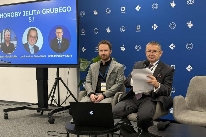 Dwóch prowadzących siedzi na krzesłach obok ekranu z planszą „Choroby Jelita Grubego 5.1”; jeden z nich trzyma mikrofon i notatki, a za nimi widać tło z logo organizatorów