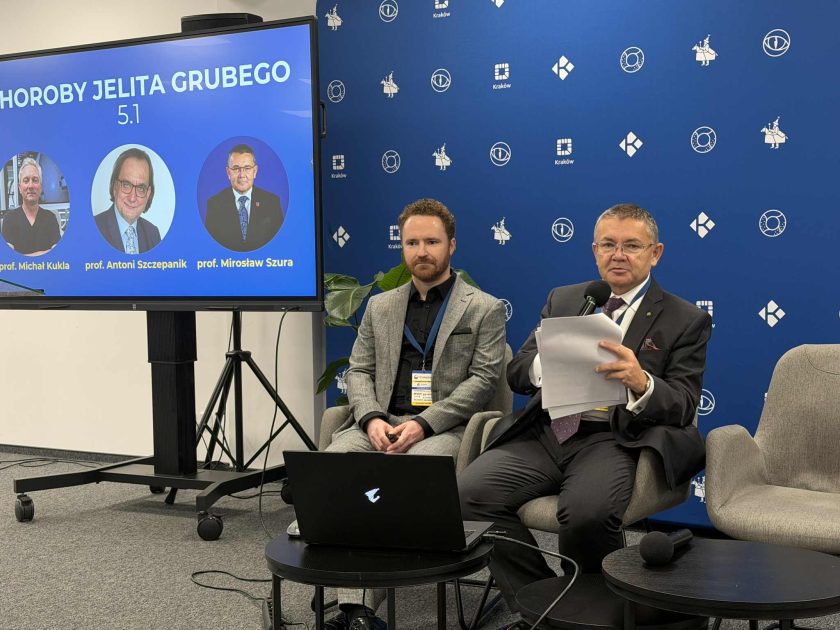 Dwóch prowadzących siedzi na krzesłach obok ekranu z planszą „Choroby Jelita Grubego 5.1”; jeden z nich trzyma mikrofon i notatki, a za nimi widać tło z logo organizatorów