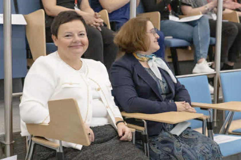 Dwie kobiety siedzą w pierwszym rzędzie w sali audytoryjnej. Kobieta po lewej, ubrana w białą marynarkę, uśmiecha się w stronę aparatu. Obok niej siedzi kobieta w granatowej marynarce i jasnej apaszce, patrząca w bok. W tle widać uczestników wydarzenia siedzących na wyższych rzędach