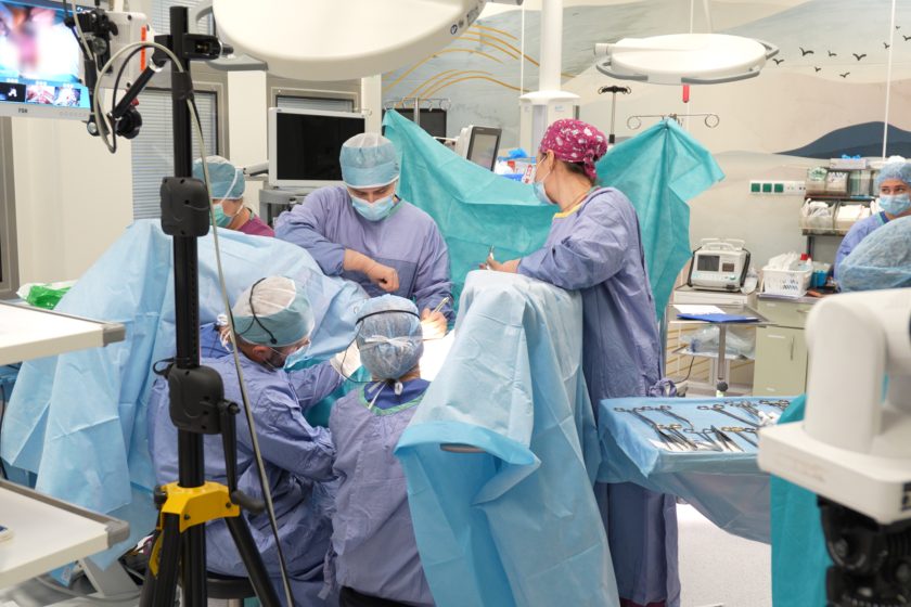 Grupa chirurgów w niebieskich fartuchach i maskach pracuje przy stole operacyjnym, otoczona sprzętem medycznym i monitorami w nowoczesnej sali zabiegowej