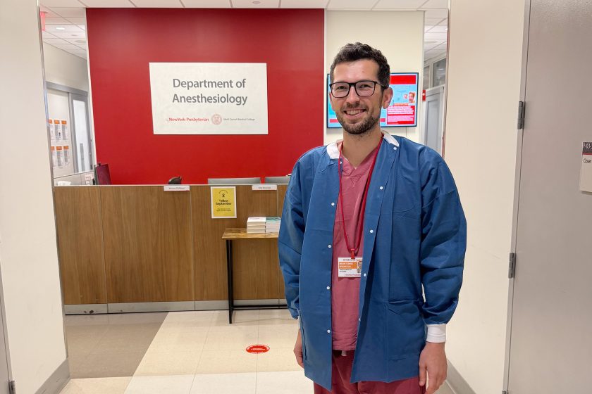 Mężczyzna w okularach, ubrany w różowy strój medyczny i niebieski fartuch ochronny, stoi w jasnym szpitalnym korytarzu przed ladą recepcyjną. Za nim widoczna jest czerwona ściana z napisem ‘Department of Anesthesiology’. Mężczyzna uśmiecha się do obiektywu