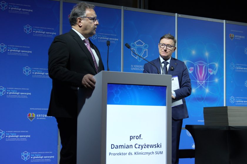 Mężczyzna w garniturze przemawia przy mównicy podczas kongresu ginekologii operacyjnej, obok stoi inny prelegent w garniturze i okularach, na tle niebieskich paneli konferencyjnych