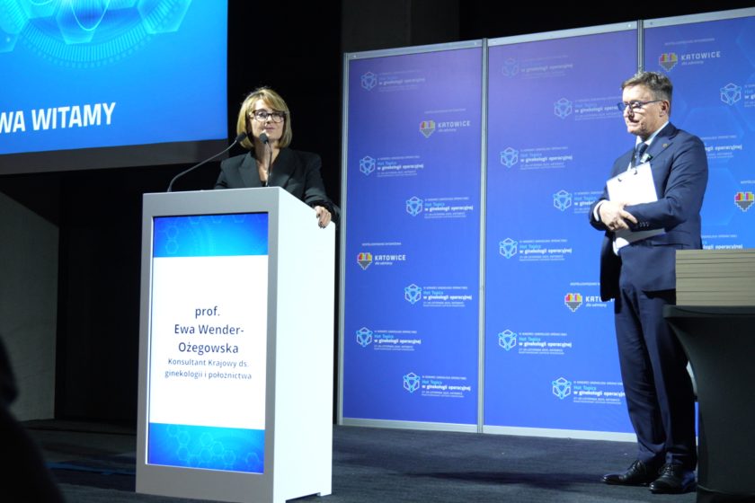 Kobieta w okularach przemawia przy mównicy, na ekranie mównicy wyświetlane jest jej nazwisko; obok niej stoi mężczyzna w garniturze trzymający dokumenty