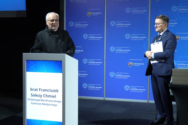 Brat zakonny w czarnym habicie przemawia z mównicy podczas kongresu, a obok niego prelegent w garniturze trzyma dokumenty i obserwuje wystąpienie