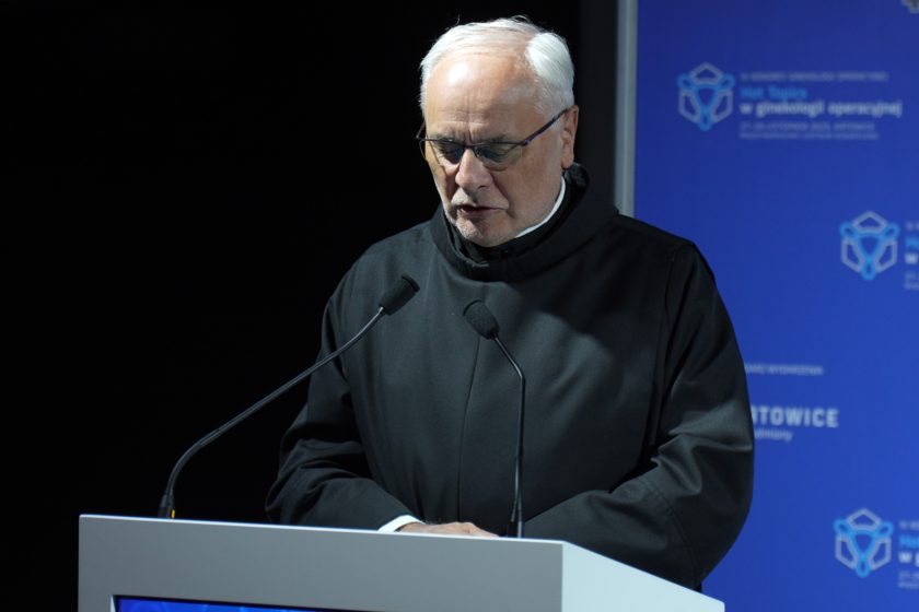 Brat zakonny w czarnym habicie stoi przy mównicy i wygłasza przemówienie, patrząc w dół na notatki, na tle niebieskiej ścianki kongresowej