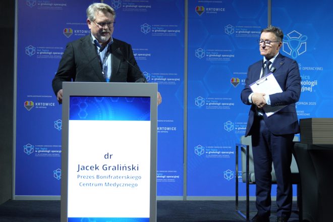 Mężczyzna w okularach i ciemnej marynarce przemawia z mównicy podczas kongresu, a obok niego stoi prelegent w garniturze trzymający teczkę i dokumenty