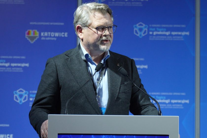 Dr Jacek Graliński przemawia przy mównicy, patrząc w bok, z dwoma mikrofonami przed sobą, na tle niebieskiej ścianki kongresowej