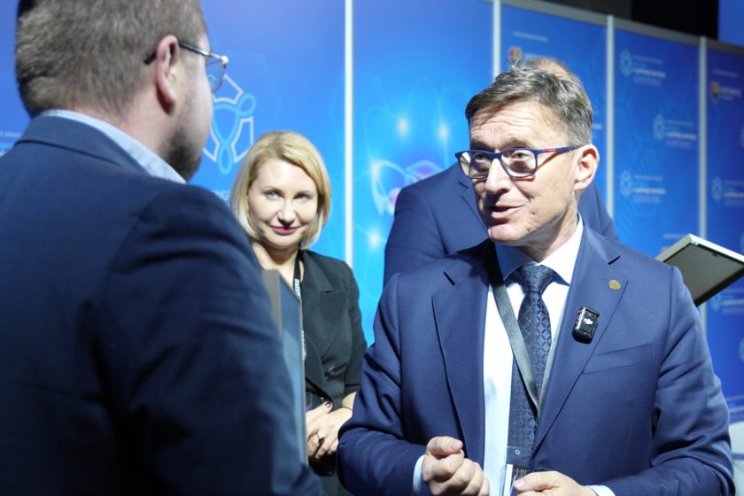 Mężczyzna w garniturze rozmawia z prelegentem w okularach, podczas gdy w tle kobieta z uśmiechem obserwuje rozmowę na tle niebieskich paneli kongresowych