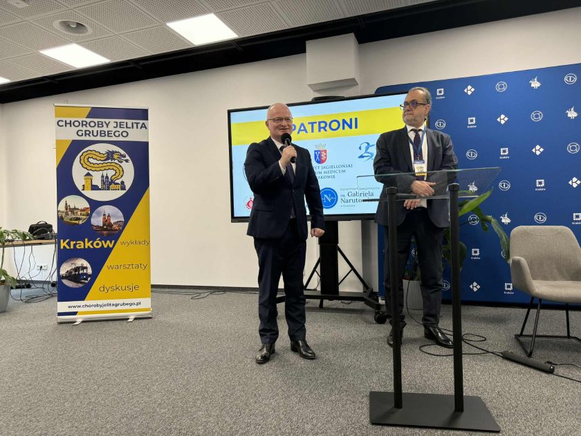 Dwóch prelegentów stoi na scenie podczas wystąpienia, obok banera konferencji oraz ekranu pokazującego logotypy patronów wydarzenia