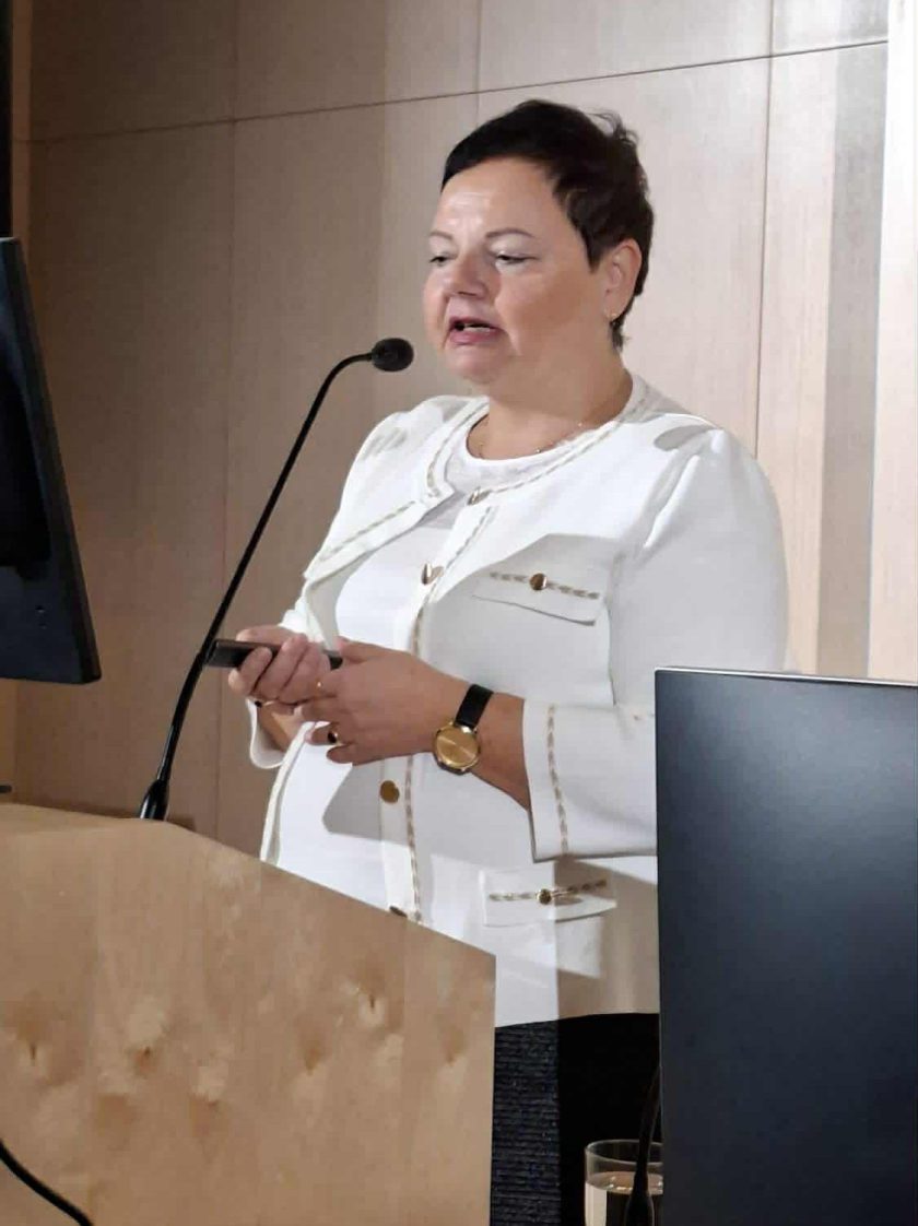 Kobieta w białej marynarce stoi przy mównicy i przemawia do mikrofonu. W dłoni trzyma pilot do prezentacji. Za nią znajduje się jasna, drewniana ściana oraz ekran prezentacyjny poza kadrem