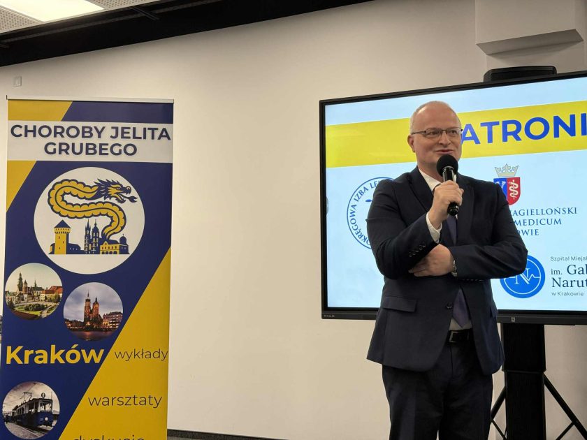 Prelegent w garniturze przemawia do mikrofonu na tle ekranu z logotypami patronów oraz banera „Choroby Jelita Grubego”