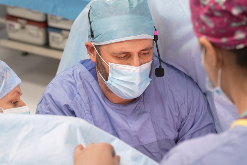 Chirurg w maseczce i czepku z założonym mikrofonem nagłownym pochyla się nad polem operacyjnym, koncentrując się na wykonywanym zabiegu