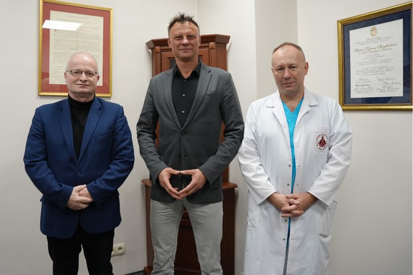 Wspólna fotografia trzech mężczyzn: przedstawiciela kierownictwa szpitala, gościa oraz lekarza w białym kitlu. Zdjęcie wykonano w gabinecie, na tle dokumentów potwierdzających tradycję i działalność jednostki