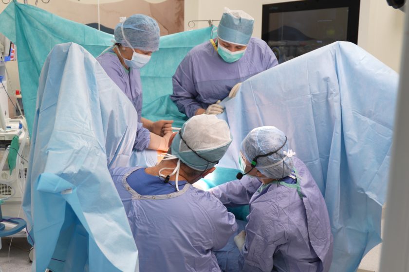 Kilku chirurgów i personel medyczny w sterylnych strojach pracuje wspólnie przy stole operacyjnym, otoczonych parawanami i sprzętem medycznym