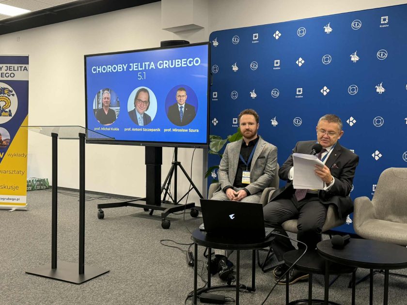 Dwóch prelegentów siedzi obok siebie przy stoliku, przed nimi laptop; za nimi ekran z grafiką „Choroby Jelita Grubego 5.1” prezentującą zdjęcia trzech specjalistów
