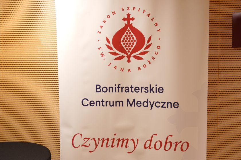 Baner z logo Zakonu Szpitalnego św. Jana Bożego oraz napisem „Bonifraterskie Centrum Medyczne – Czynimy dobro”, ustawiony w pomieszczeniu konferencyjnym