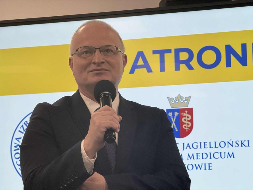 Zbliżenie na mężczyznę przemawiającego do mikrofonu, stojącego na tle ekranu prezentującego logotypy patronów konferencji