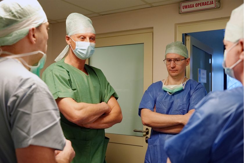 Lekarze w strojach operacyjnych omawiają szczegóły procedury przed wejściem na salę zabiegową. Na drzwiach widnieje ostrzegawczy napis „Uwaga operacja”