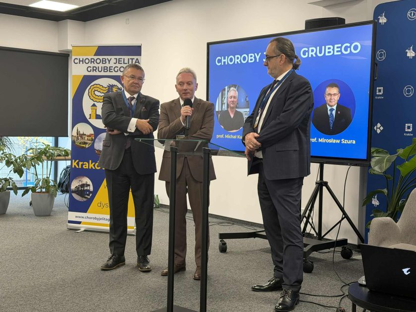 Trzech prelegentów prowadzi wspólne wystąpienie, stojąc przy przezroczystej mównicy; w tle ekran z nazwiskami ekspertów biorących udział w wydarzeniu