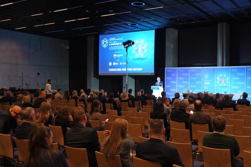 Uczestnicy kongresu siedzą w rzędach krzeseł w dużej sali konferencyjnej, a na scenie prelegent przemawia przy mównicy obok dużego ekranu