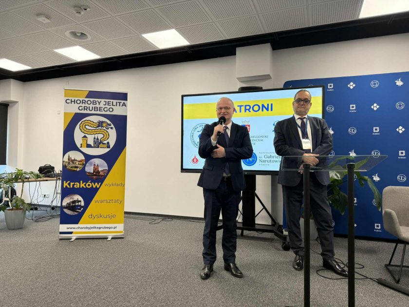 Dwóch prelegentów w garniturach stoi przy mównicy i prowadzi wystąpienie; obok znajduje się baner wydarzenia oraz ekran z logotypami patronów konferencji