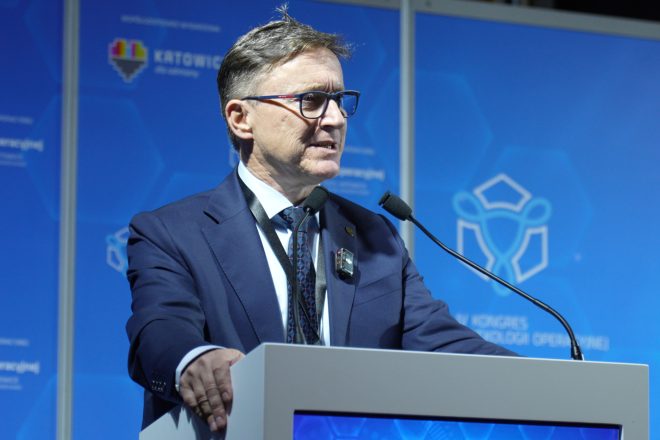 Mężczyzna w garniturze i okularach przemawia przy mównicy na tle niebieskiej ścianki kongresu, wyposażony w mikrofony i identyfikator