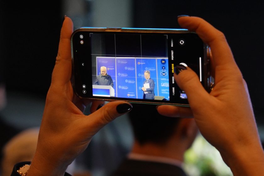 Zbliżenie na dłonie trzymające smartfon, na którego ekranie widać nagrywaną scenę z dwoma prelegentami stojącymi przy mównicy na tle ścianek konferencyjnych