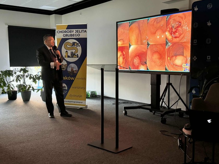 Prelegent w garniturze omawia obrazy endoskopowe jelita grubego wyświetlone na dużym ekranie; po lewej stronie widoczny baner konferencji