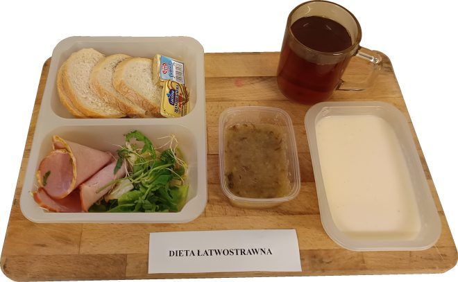 Taca ze śniadaniem w diecie łatwostrawnej. Na tacy znajduje się kilka kromek białego pieczywa, masło i serek topiony, plasterki wędliny z listkami sałaty, porcje musu owocowego i kremowej zupy mlecznej oraz szklanka herbaty. Posiłek ułożony w plastikowych pojemnikach na drewnianej tacy z napisem „Dieta łatwostrawna”