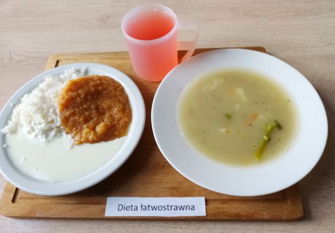 Na drewnianej tacy znajduje się zestaw obiadowy w diecie łatwostrawnej. Na jednym talerzu podano porcję białego ryżu polanego jasnym sosem i podanego z musem z duszonych jabłek lub marchwi. Obok stoi talerz z zupą jarzynową o delikatnej konsystencji, w której widać kawałki brokułów i marchewki. Do zestawu dołączony jest napój o różowym kolorze w przezroczystym kubku. Przed tacą leży kartka z napisem „Dieta łatwostrawna”