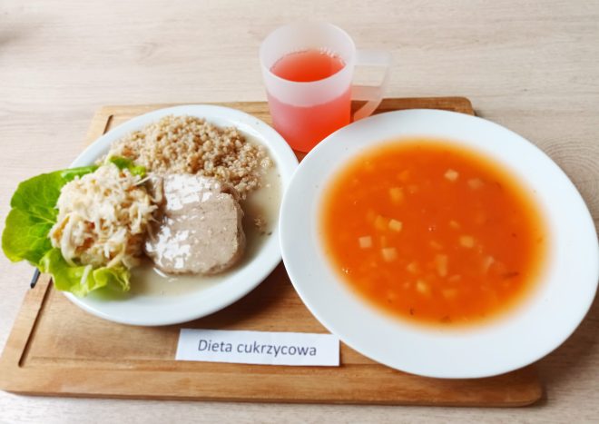 Na drewnianej tacy ustawiony jest posiłek oznaczony etykietą „Dieta cukrzycowa”. Na talerzu znajduje się porcja kaszy gryczanej, dwa plasterki duszonego mięsa w jasnym sosie, oraz surówka z białej kapusty i marchewki ułożona na liściu sałaty. Obok talerza stoi miska z zupą pomidorową z kawałkami ziemniaków oraz kubek z czerwonym napojem