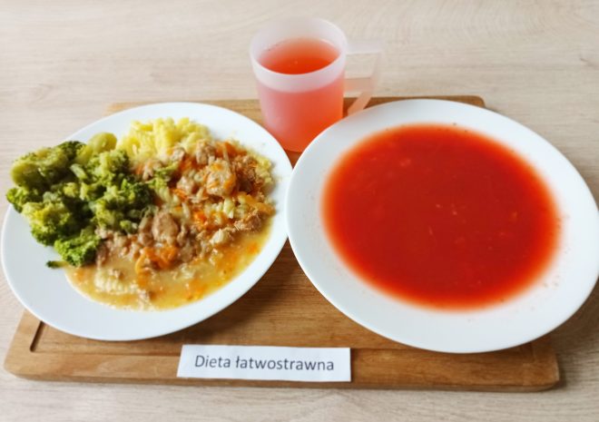 Na tacy dwa talerze: na jednym makaron z sosem warzywno-mięsnym i gotowanymi brokułami, na drugim zupa pomidorowa o gładkiej konsystencji. Do posiłku podany napój owocowy w przezroczystym kubku. Przed tacką kartka z napisem „Dieta łatwostrawna”