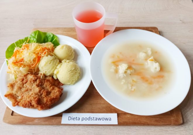 Na tacy ustawiony talerz z panierowanym kotletem schabowym, trzema porcjami ziemniaków z koperkiem i surówką z kapusty kiszonej z marchewką. Obok drugi talerz z zupą kalafiorową z marchewką. W przezroczystym kubku napój o czerwonym kolorze. Przed tacą karteczka z napisem „Dieta podstawowa”