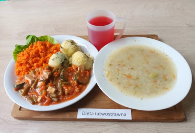 Na tacy z napisem „Dieta łatwostrawna” podano obiad: na dużym talerzu znajduje się gęsta zupa jarzynowa z ryżem, a obok – talerz z drugim daniem, na którym widać porcję gotowanych ziemniaków z koperkiem, duszone warzywa w sosie pomidorowym z kawałkami mięsa oraz surówkę z drobno startego gotowanego buraka lub marchwi. Do posiłku podano kubek z czerwonym napojem owocowym