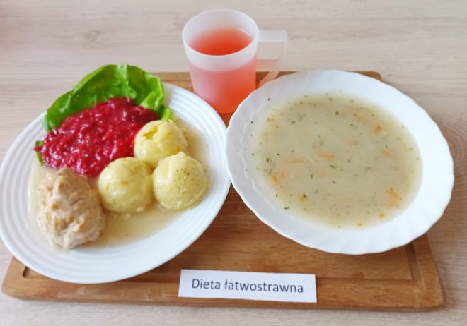 Na drewnianej tacy talerz z pieczywem pszennym i bułką, plasterkami wędliny drobiowej, twarożkiem, masłem, pomidorem, dżemem i jabłkiem. Obok miseczka z mlekiem i napój w przezroczystym kubku. Na tacy widoczna karteczka z napisem „Dieta łatwostrawna”