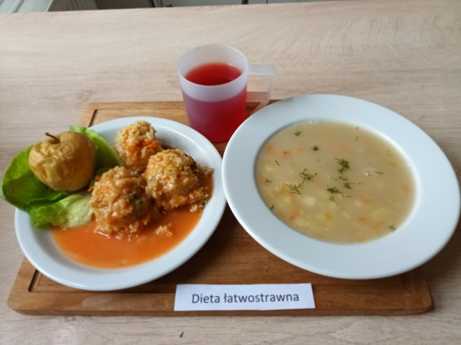 Zestaw obiadowy w diecie łatwostrawnej: talerz z jasną zupą jarzynową posypaną koperkiem oraz talerz z pulpetami w sosie pomidorowym, podanymi z pieczonym jabłkiem na liściu sałaty. Obok kubek czerwonego kompotu