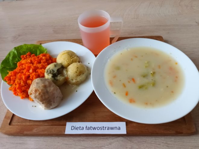 Obiad w ramach „Diety łatwostrawnej”. Na talerzu podano gotowane marchewki, liść sałaty, kulki ziemniaczane z ziołami oraz pulpet w delikatnym sosie. Obok znajduje się talerz z jasną zupą warzywną, a w tle kubek z różowym napojem