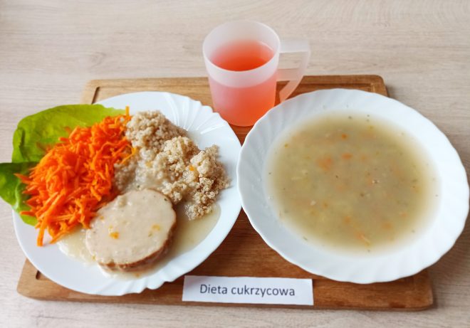Posiłek składa się z talerza z tartą marchewką, kaszą oraz plasterkiem mięsa w sosie. Obok znajduje się miseczka z zupą jarzynową oraz kubek z różowym napojem. Na tacy widoczna jest etykieta „Dieta cukrzycowa”