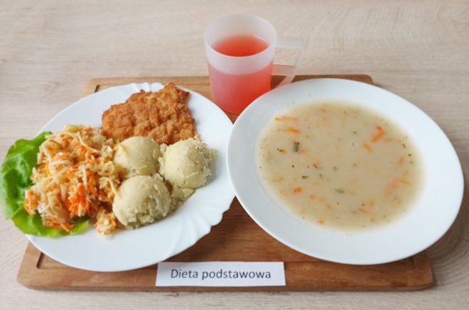 Talerz z kotletem panierowanym, porcją surówki z marchewki i białej kapusty na liściu sałaty oraz trzema kulkami ziemniaków. Obok miska z jasną zupą jarzynową i kubek z czerwonym kompotem. Pod zestawem napis ‘Dieta podstawowa‘