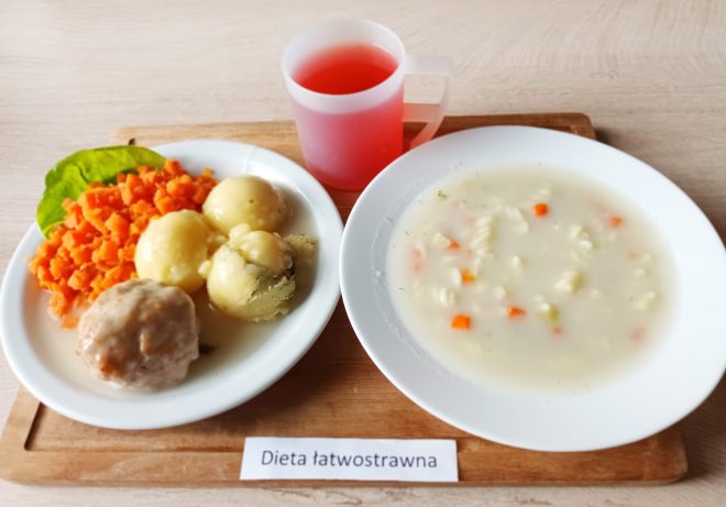 Zestaw obiadowy składający się z pulpetu w białym sosie, gotowanych ziemniaków, drobno pokrojonej gotowanej marchewki oraz talerza z jasną zupą z makaronem i warzywami. Obok ustawiony jest kubek z czerwonym napojem. Przed posiłkiem widoczna etykieta z napisem „Dieta łatwostrawna”