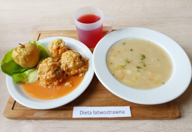 Zestaw obiadowy w diecie łatwostrawnej: talerz z zupą jarzynową o kremowej konsystencji, obok talerz z pulpetami w lekkim sosie pomidorowym, podanymi na liściach sałaty z pieczonym jabłkiem. Do tego kubek czerwonego kompotu. Całość ustawiona na drewnianej tacy z podpisem „Dieta łatwostrawna”