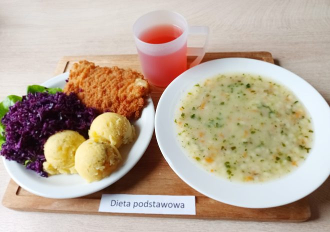 Obiad w diecie podstawowej: talerz z kotletem panierowanym, dwoma kulkami ziemniaków oraz porcją surówki z czerwonej kapusty. Obok talerz z zupą jarzynową. Do posiłku podany różowy kompot. Całość na drewnianej tacy oznaczonej „Dieta podstawowa”