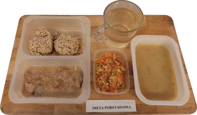 Taca z obiadem w diecie podstawowej. W zestawie znajduje się porcja kaszy jęczmiennej, gulasz z kawałkami mięsa w sosie, surówka z kapusty i marchewki, zupa krem w jasnobrązowym kolorze oraz szklanka kompotu lub herbaty owocowej. Posiłek podany w plastikowych pojemnikach na drewnianej tacy z napisem „Dieta podstawowa”