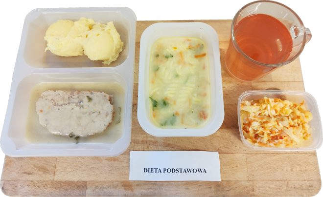 Na zdjęciu widoczny zestaw obiadowy w ramach diety podstawowej, podany na drewnianej tacy. W plastikowych pojemnikach znajdują się: zupa jarzynowa z makaronem, ziemniaki puree z kotletem mielonym w sosie oraz surówka z kapusty i marchwi. Do posiłku dołączony jest kubek z czerwonawym kompotem. Przed zestawem leży kartka z napisem „DIETA PODSTAWOWA”